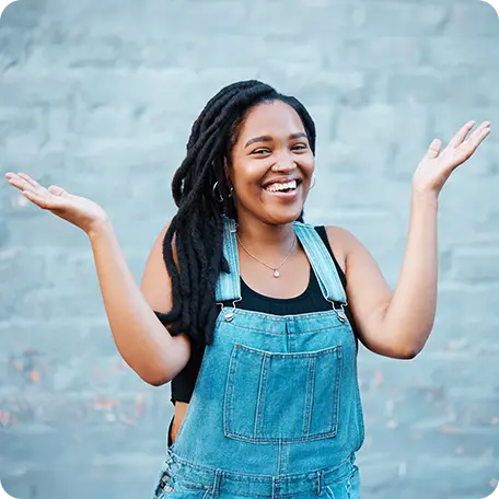 Smiling young woman with dreadlocks wearing denim overalls, holding her hands out in a welcoming gesture against a blue wall, symbolizing acceptance and relief.