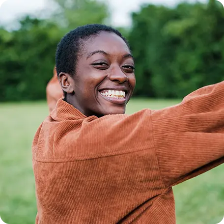 Smiling young woman with short dark hair and a brown shirt, standing in a field with arms slightly outstretched, conveying openness and happiness.