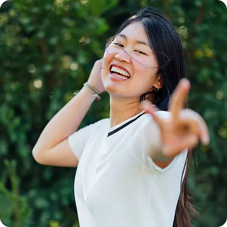 Smiling young woman with long dark hair holding up a peace sign, with a blurred green background, expressing inner peace and well-being.