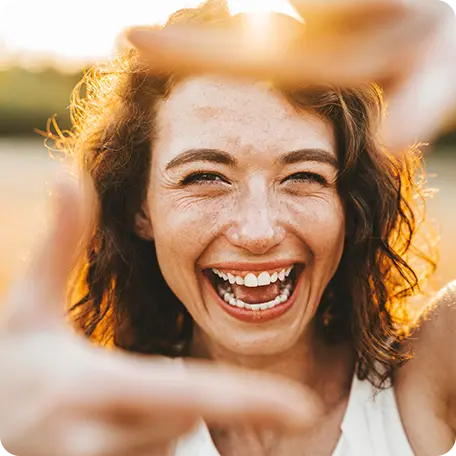 Close-up of a smiling woman with curly hair and freckles, making a frame gesture with her hands towards the camera, with bright sunlight behind her, emphasizing focus and a bright future.