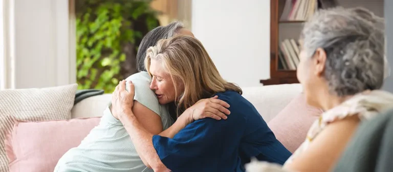 Two older women embrace in a deep, comforting hug on a sofa, symbolizing mutual emotional support and finding help during a time of grief or loss.
