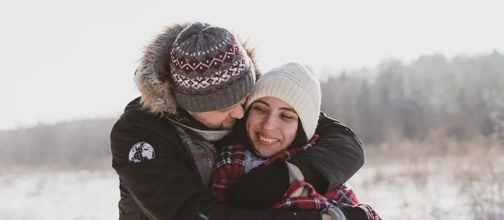 A happy couple embraces closely outdoors in the bright winter snow, both wearing warm knit hats and scarves. The man is hugging the smiling woman from behind.